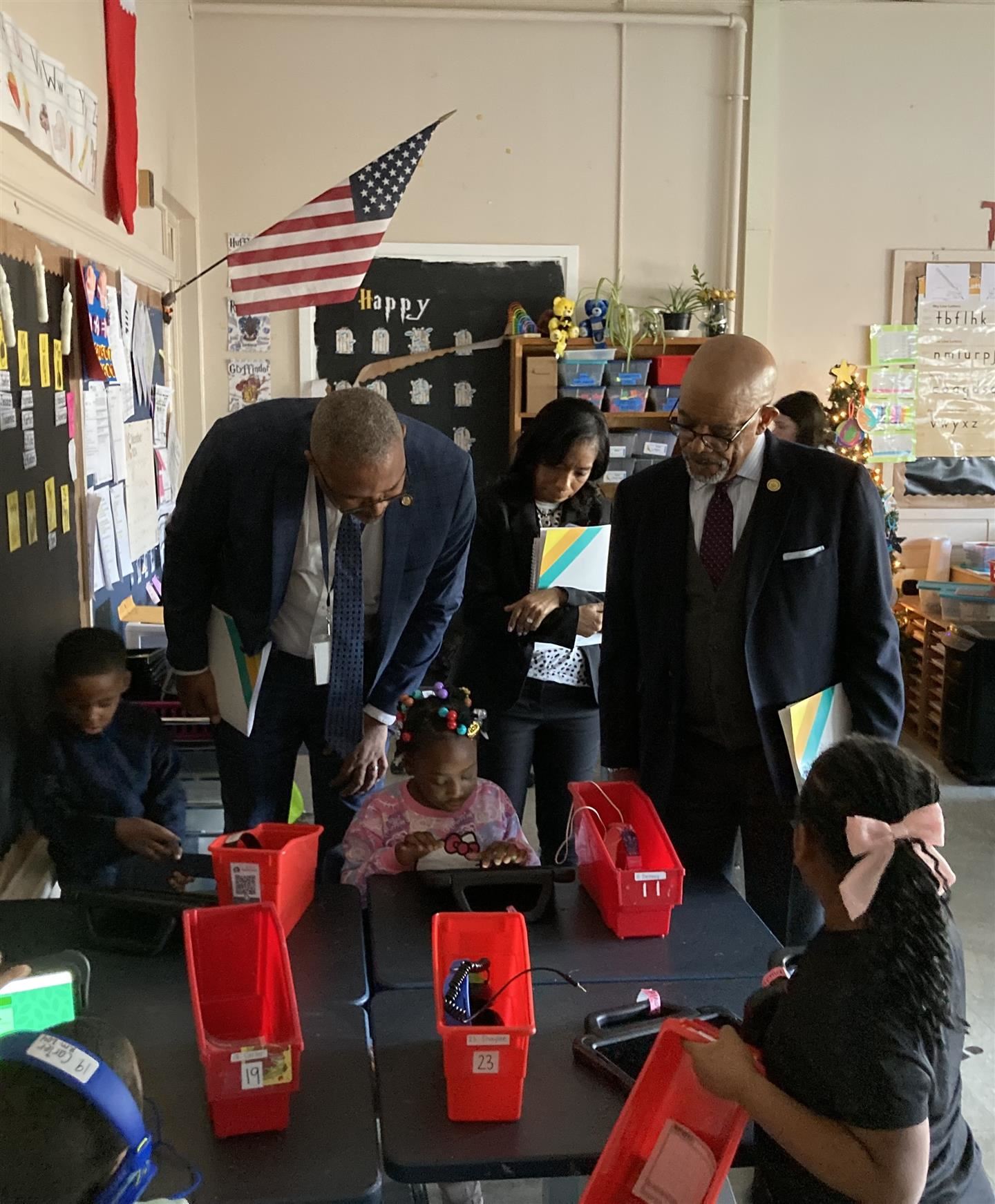 Three school leaders look over a student's shoulder helping her on a tablet.